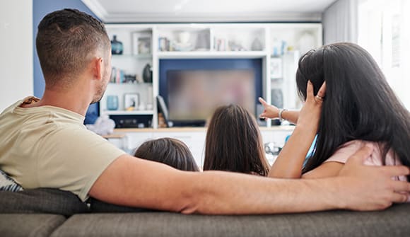 Two parents sit on a couch with two children between them, facing away from the camera watching a tv in the background that the mom is pointing to as if they are talking about it