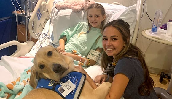 Jude lays on the bed with a patient while his handler, Child Life specialist Kara Williams, CCLS, CTRS, kneels next to the bed.