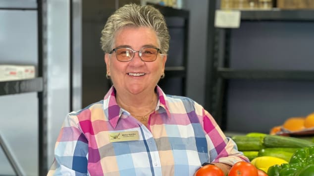 photo of a volunteer at pie in the sky smiling, standing next to fresh fruit and vegetables in a warehouse.