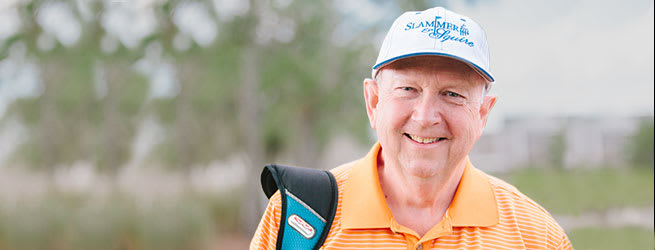 photo of patient Jim Marshall wearing an orange shirt and baseball cap standing on a golf course and carrying a golf bag on his shoulder