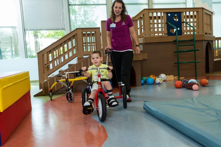 photo of a pediatric clinic setting where a female provider pushes a young male patient on a tricycle
