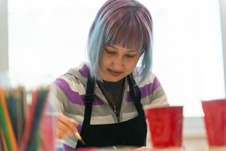 a female teenager with purple-and-blue-dyed hair wears a black smock while sitting at a table and drawing with colored pencils