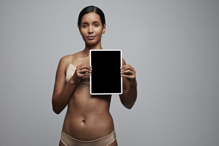 woman in underwear standing against a neutral background and holding a screen over her left breast