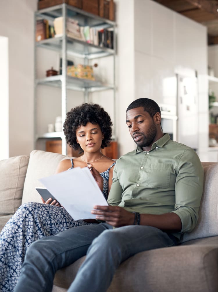 An african-american couple in their 30s sitting on a sofa of their home reading an advance directive document.
