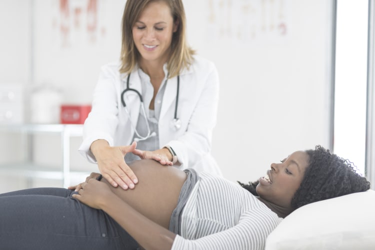 A pregnant woman is laying on an examination table at the doctor's office while a female physician exams her belly.