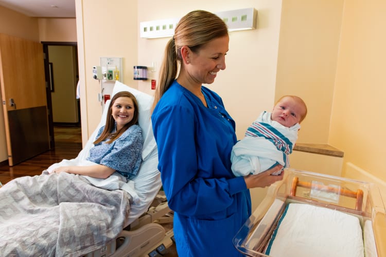 A female nurse holds a newborn baby wrapped in a blanket while the new mother looks on smiling