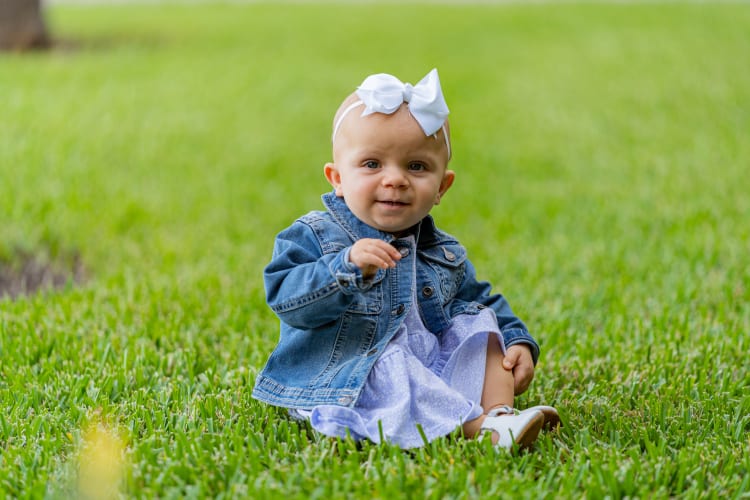 smiling baby girl who had a brain tumor removed sits in a field of grass