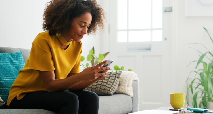 smiling woman scrolling through her phone while sitting on a couch in a brightly lit room