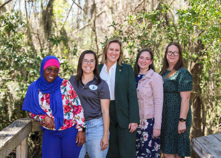 group of smiling woman of varying ages stand together on a boardwalk in the woods