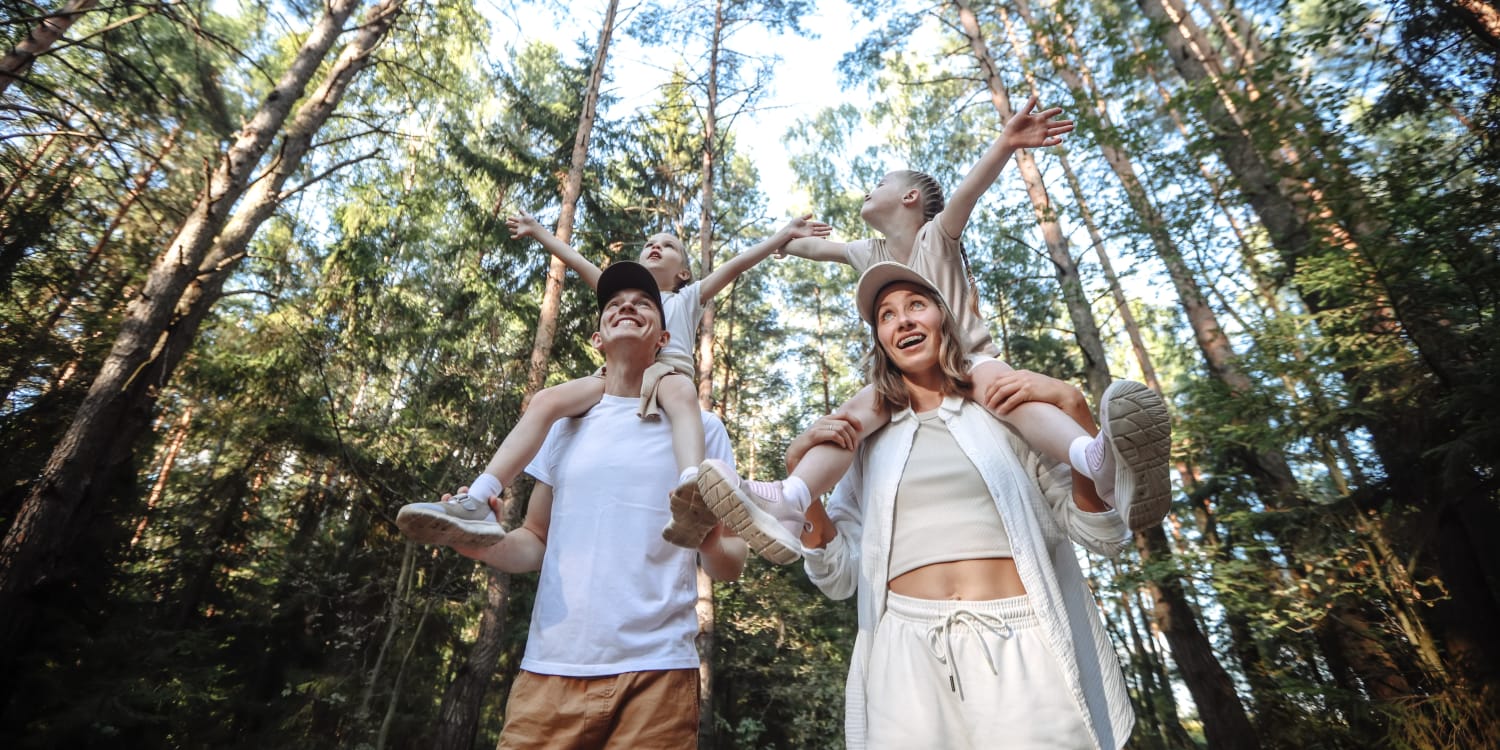 mom and dad with two school age girls on their shoulders enjoying their time in a pine tree area of their neighborhood.