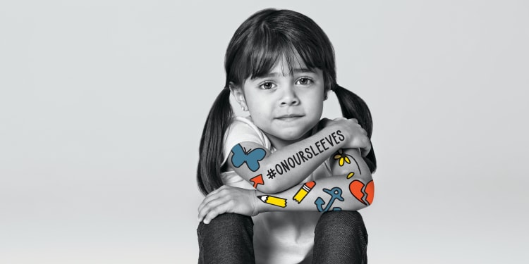 a school age child with dark hair sits, looking at the camera, and has colorful artwork on her arms, and the hashtag On Our Sleeves.