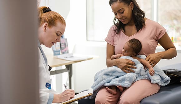 Female doctor updates a newborn's medical record as he and his mother sit on the exam table