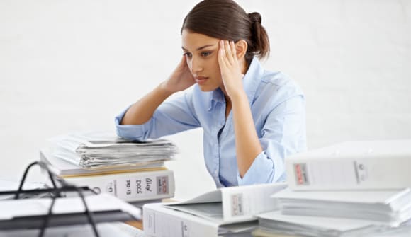 Woman looking stressed is sitting at desk and surrounded by binders and papers