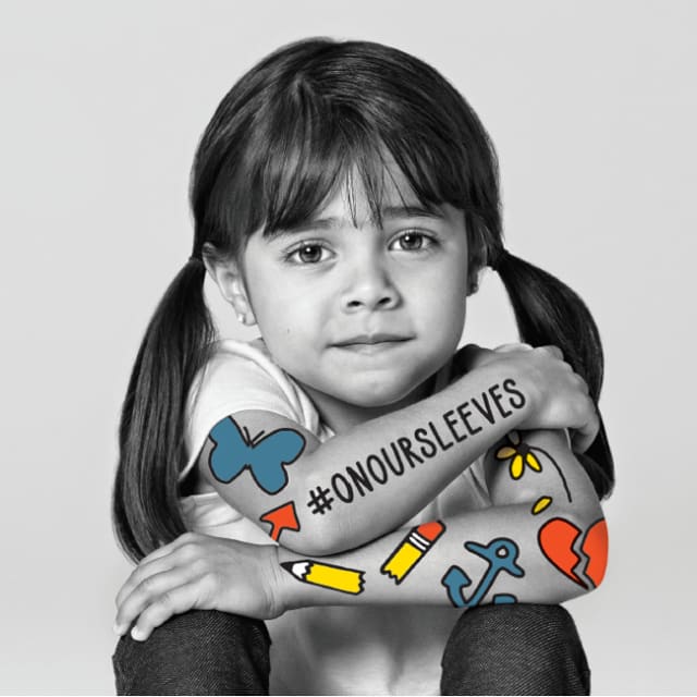 black and white photo of a young girl with pigtails looking at the camera and sitting with her arms crossed on her knees. On her arms is written #onoursleeves and graphics of a butterfly, broken pencil, anchor, broken heart, arrow, and a daisy with a petal that has fallen off.
