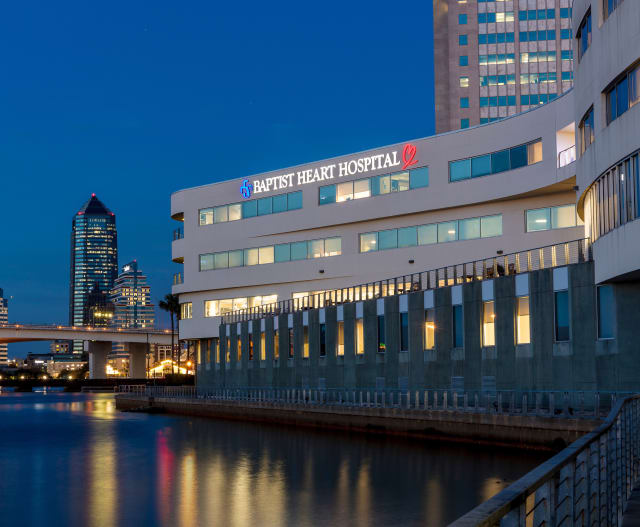 A nighttime photograph of the Baptist Heart Hospital exterior from the St. Johns River with city buildings in the background.