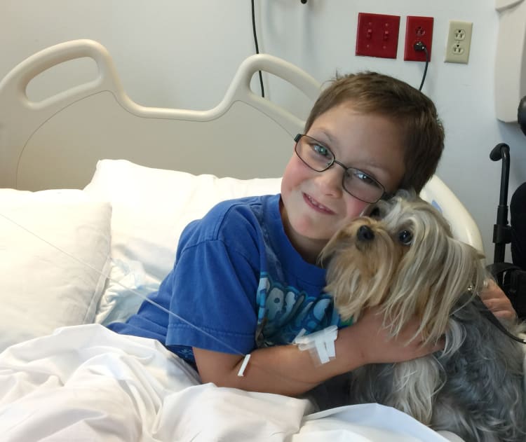 smiling young boy hugging a small dog in a hospital bed