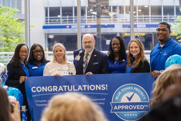 baptist health and blue zones employees standing in front of a banner that says "congratulations on being Blue Zones Project approved"