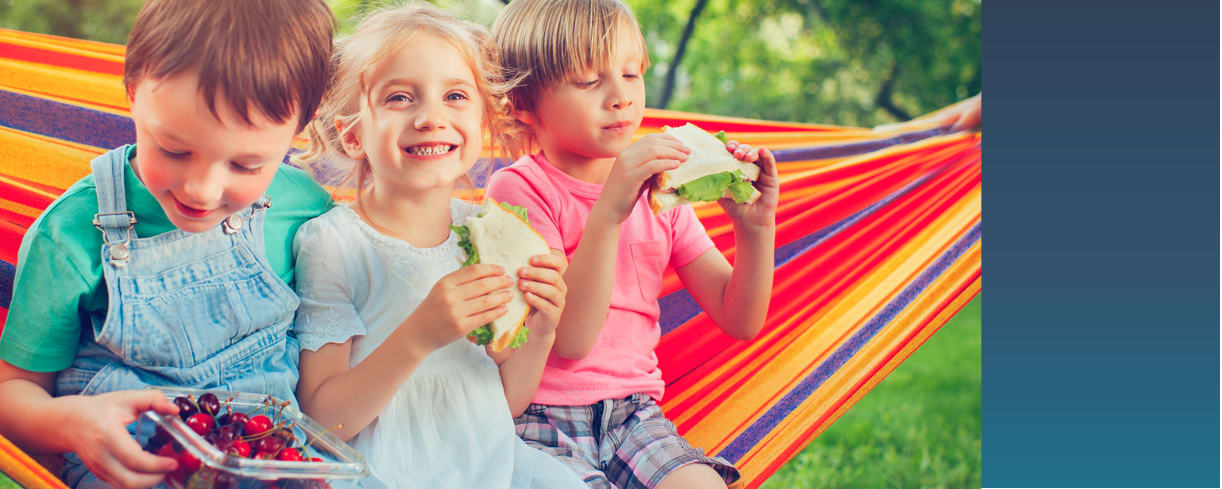 Three school-age children sitting on a striped hammock outside eating sandwiches with smiles on their faces