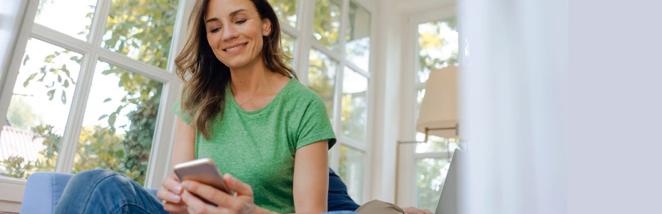 Woman in a brightly lit home looking at her smartphone while sitting casually smiling during a virtual visit using Baptist HealthPlace app.
