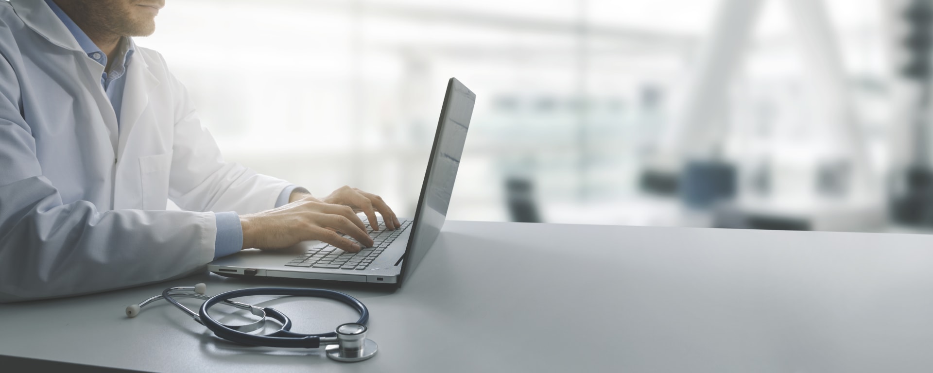 a healthcare provider typing on a laptop computer with a stethoscope placed on the table next to the laptop.