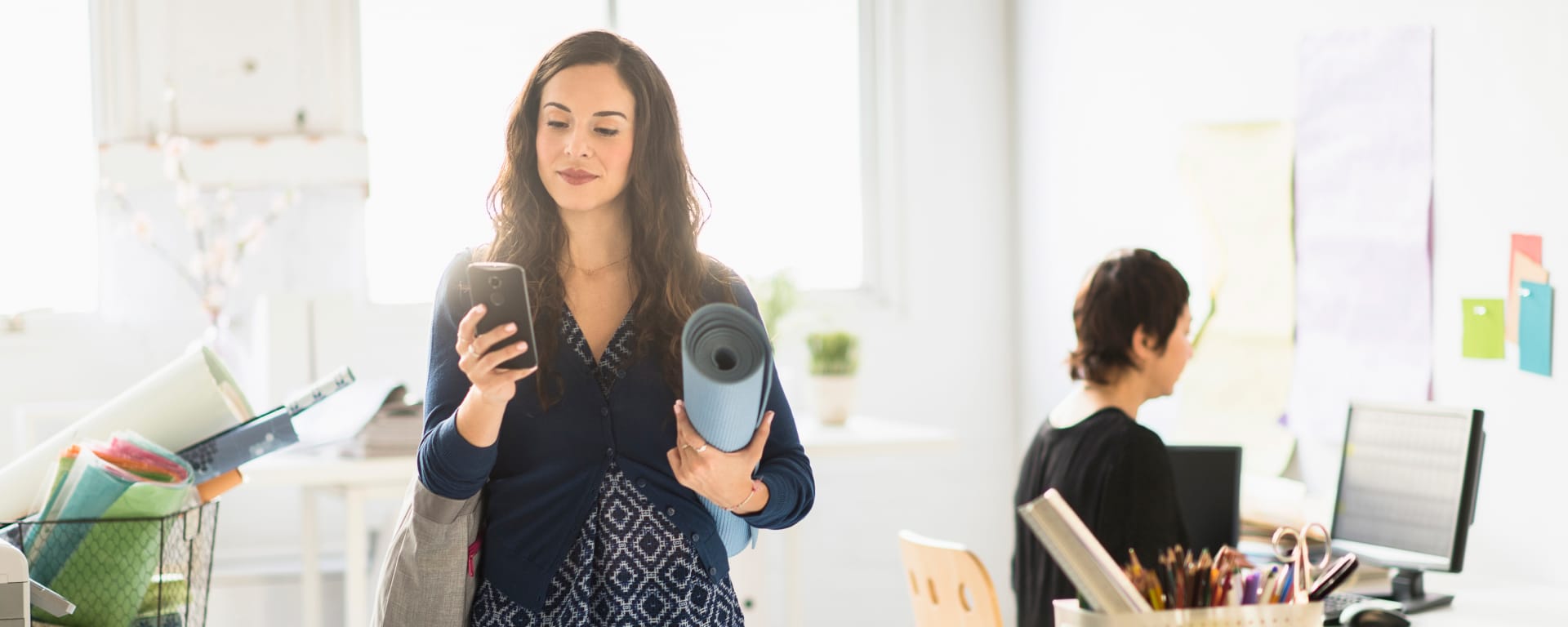 woman looking at her phone, holding a yoga mat in a brightly lit office space