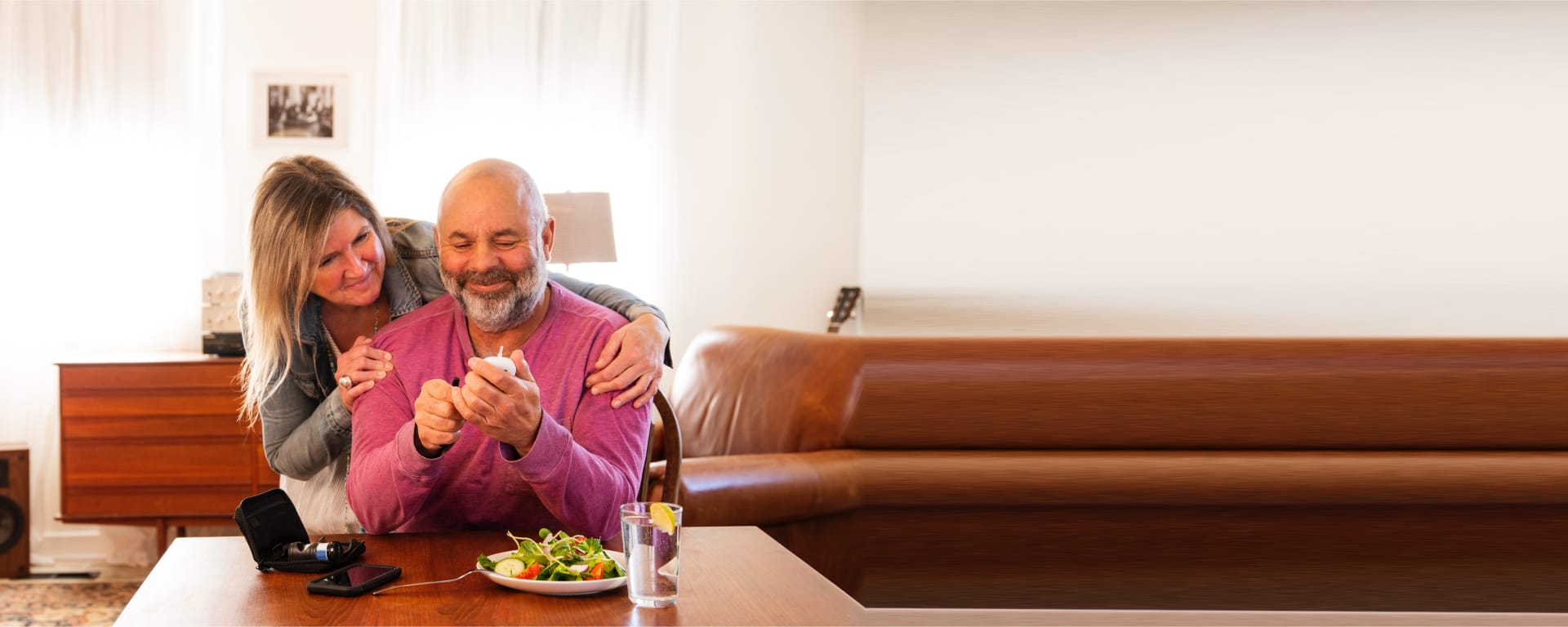 a happy man sitting at the dining table checking his blood sugar on a diabetes device while his wife looks on smiling.
