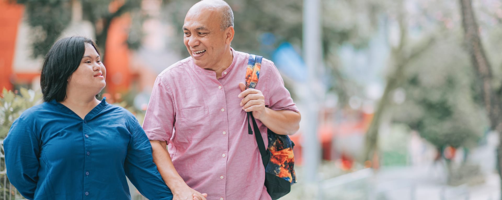 A young woman with Down syndrome holds the hand of an older man while walking down the street, both are smiling