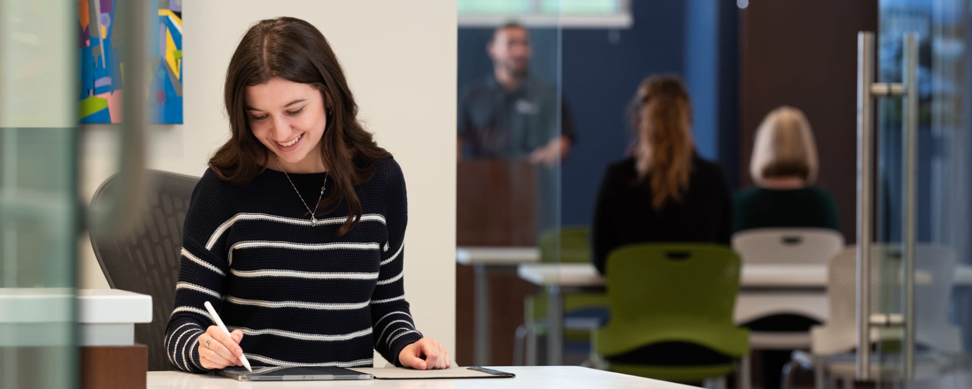woman sits at a desk while a seminar goes on behind her