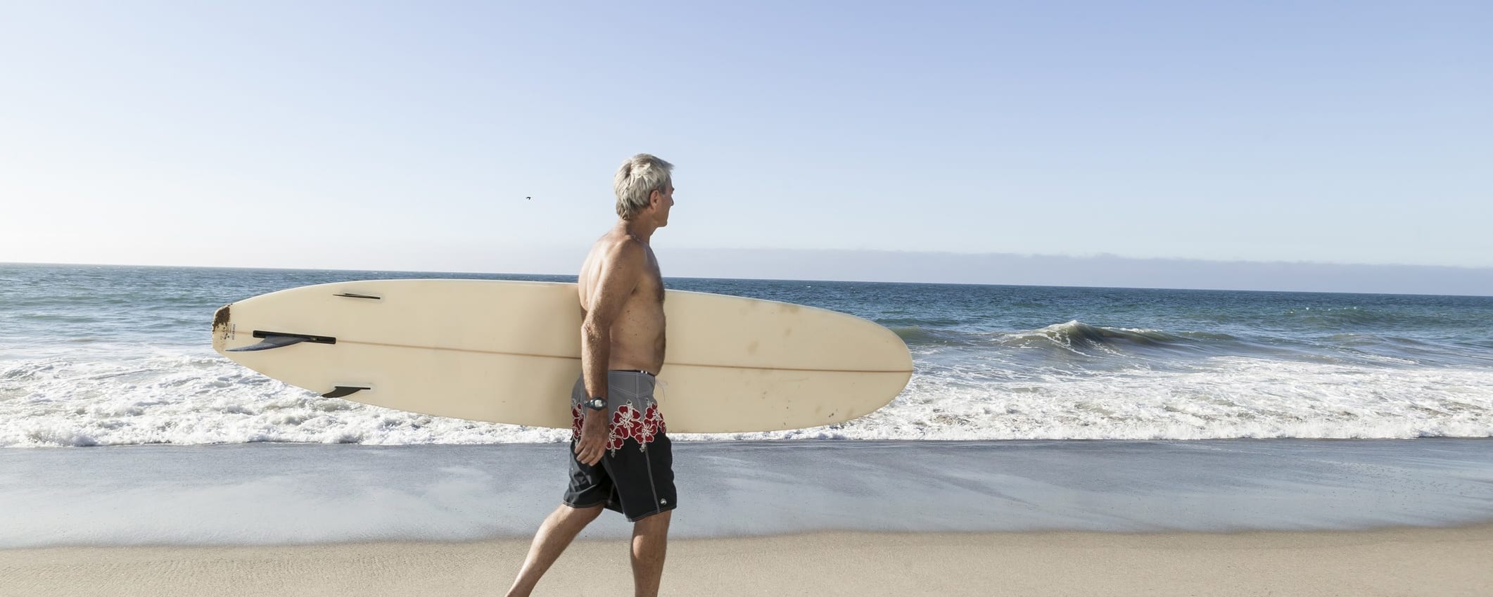 A man with gray hair walks along a beach carrying a surf board