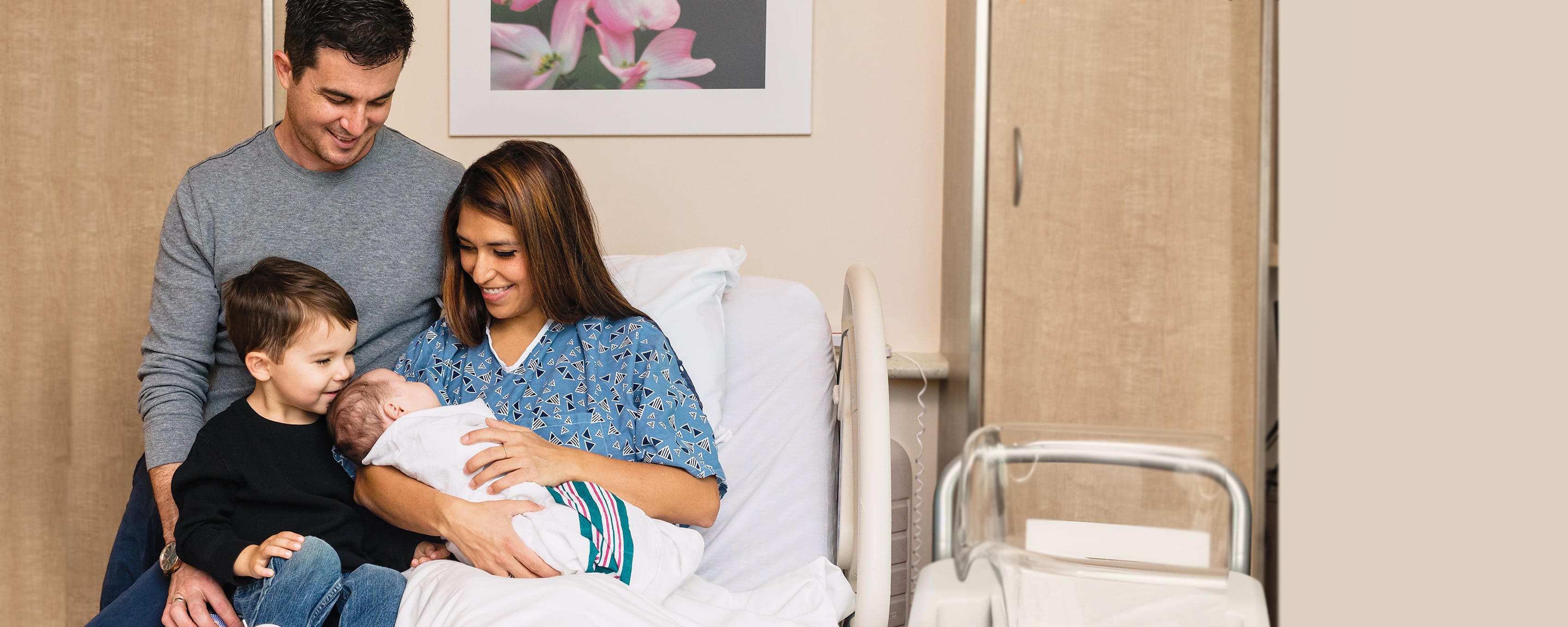 A mother holds her newborn baby wrapped in a blanket while the husband and young sibling look on