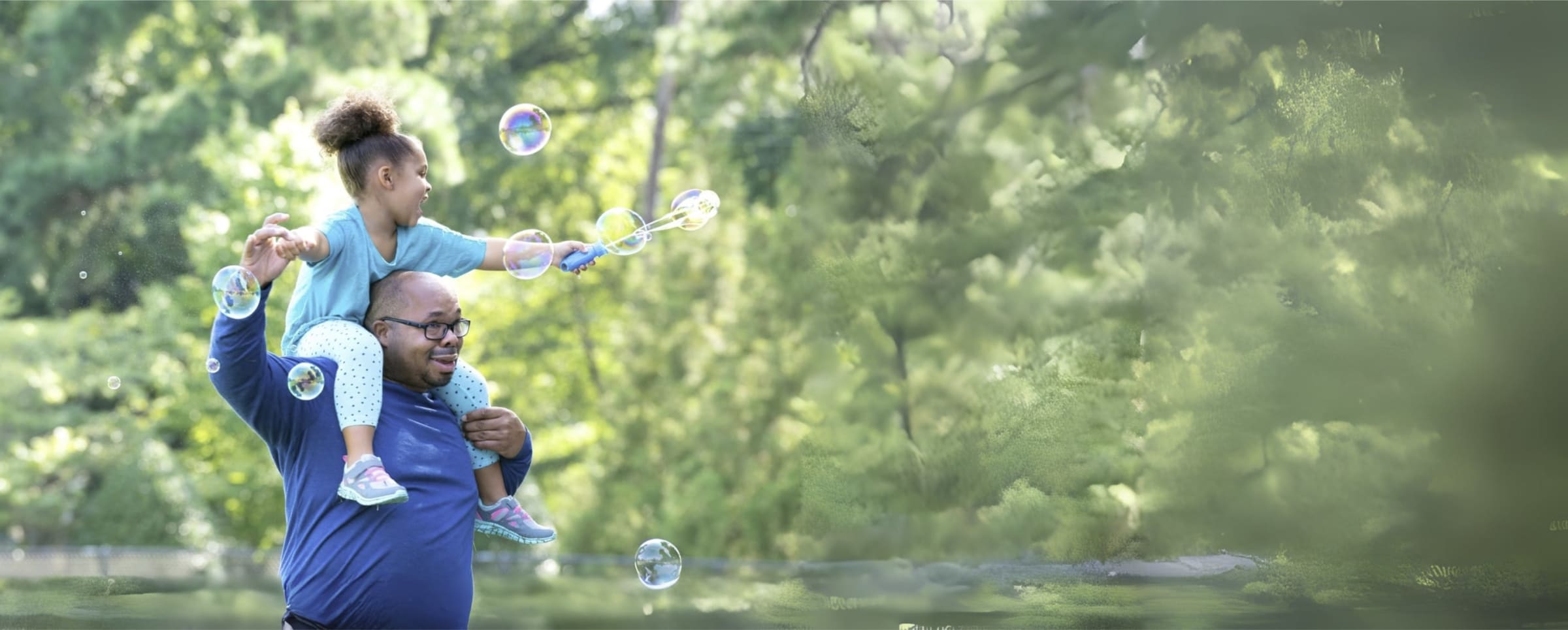 A smiling father in a park full of trees with his happy daughter on his shoulders making bubbles with a bubble wand.