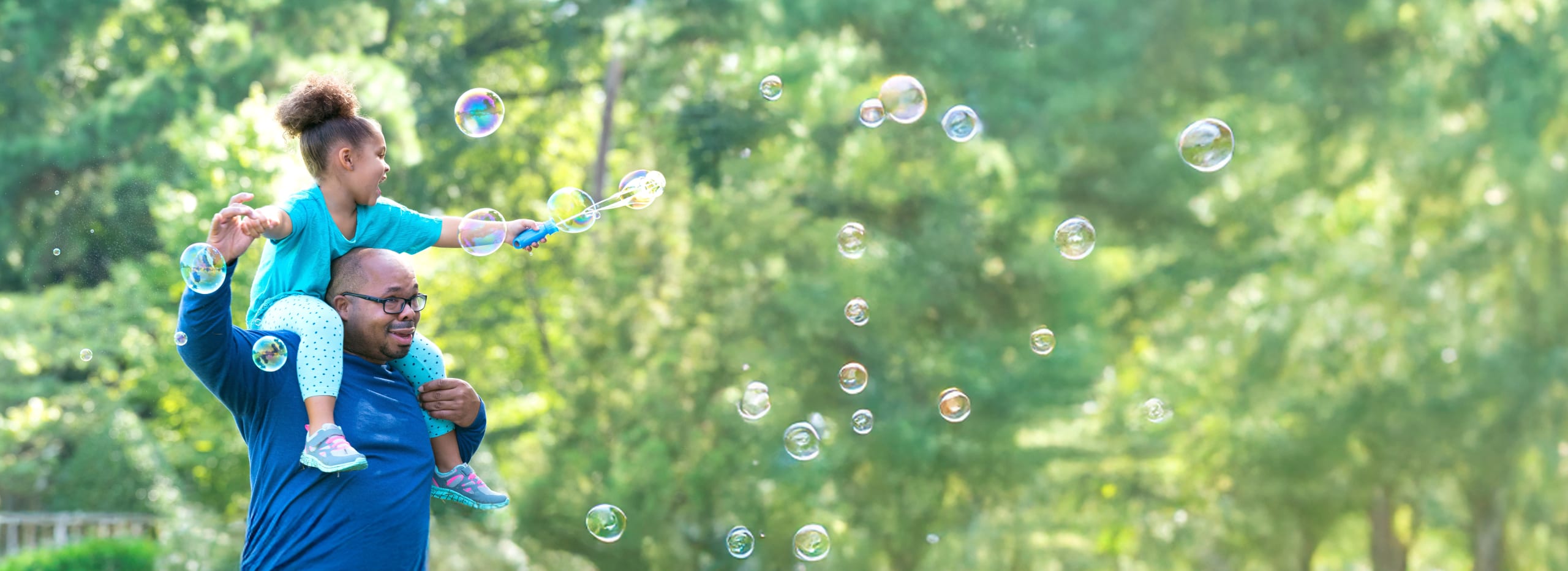 A smiling father in a park full of trees with his happy daughter on his shoulders making bubbles with a bubble wand.