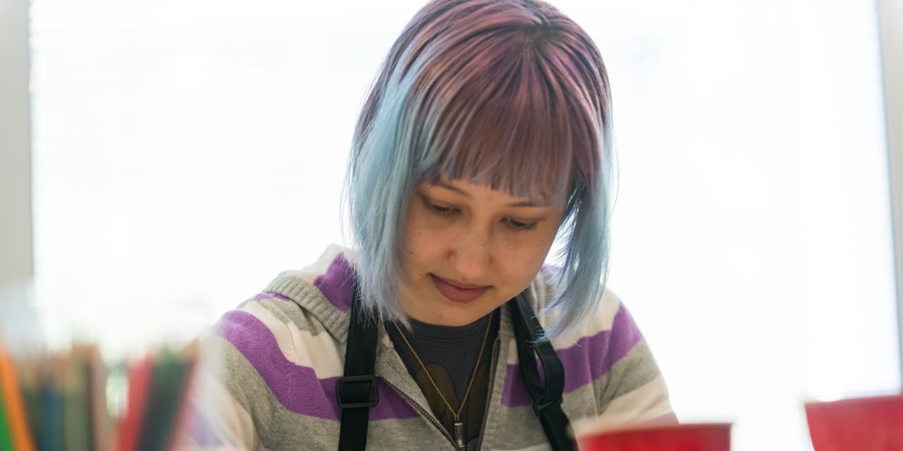 a female teenager with purple-and-blue-dyed hair wears a black smock while sitting at a table and drawing with colored pencils