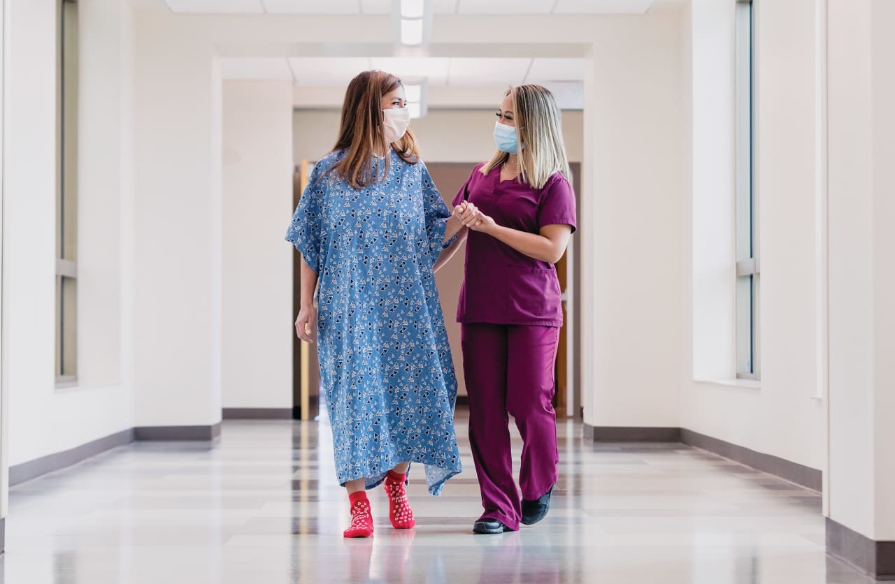 A female nurse helps a female patient walk down a hospital hallway