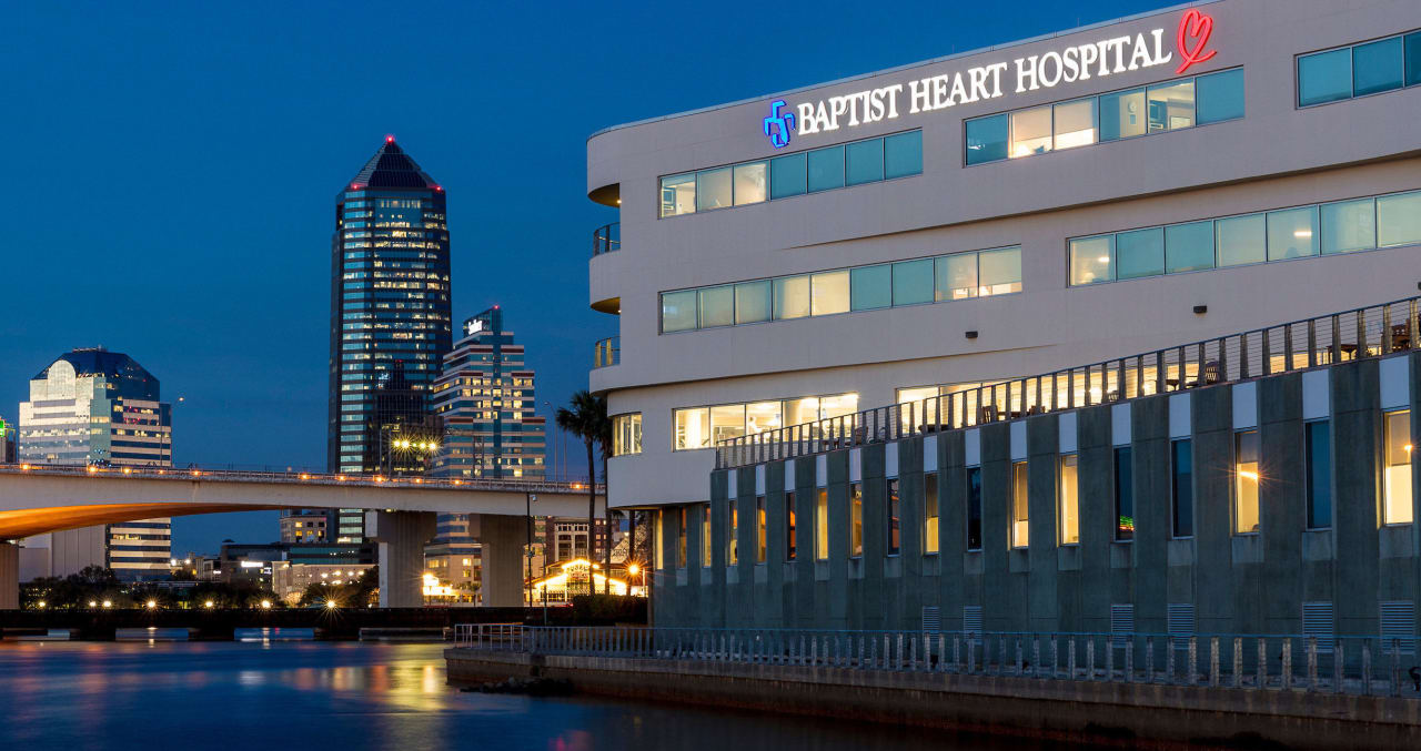 A nighttime photograph of the Baptist Heart Hospital exterior from the St. Johns River with city buildings in the background.