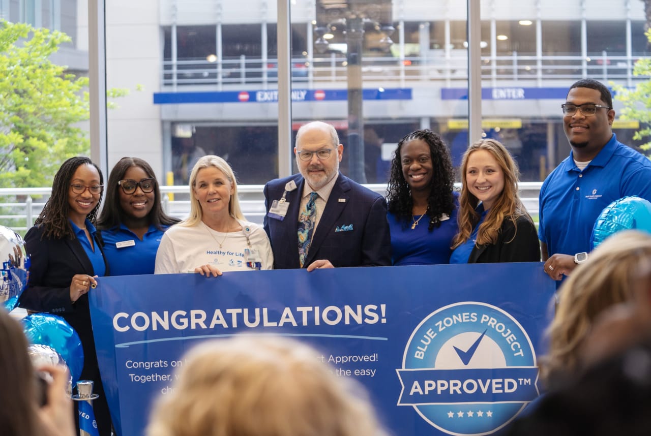 baptist health and blue zones employees standing in front of a banner that says "congratulations on being Blue Zones Project approved"
