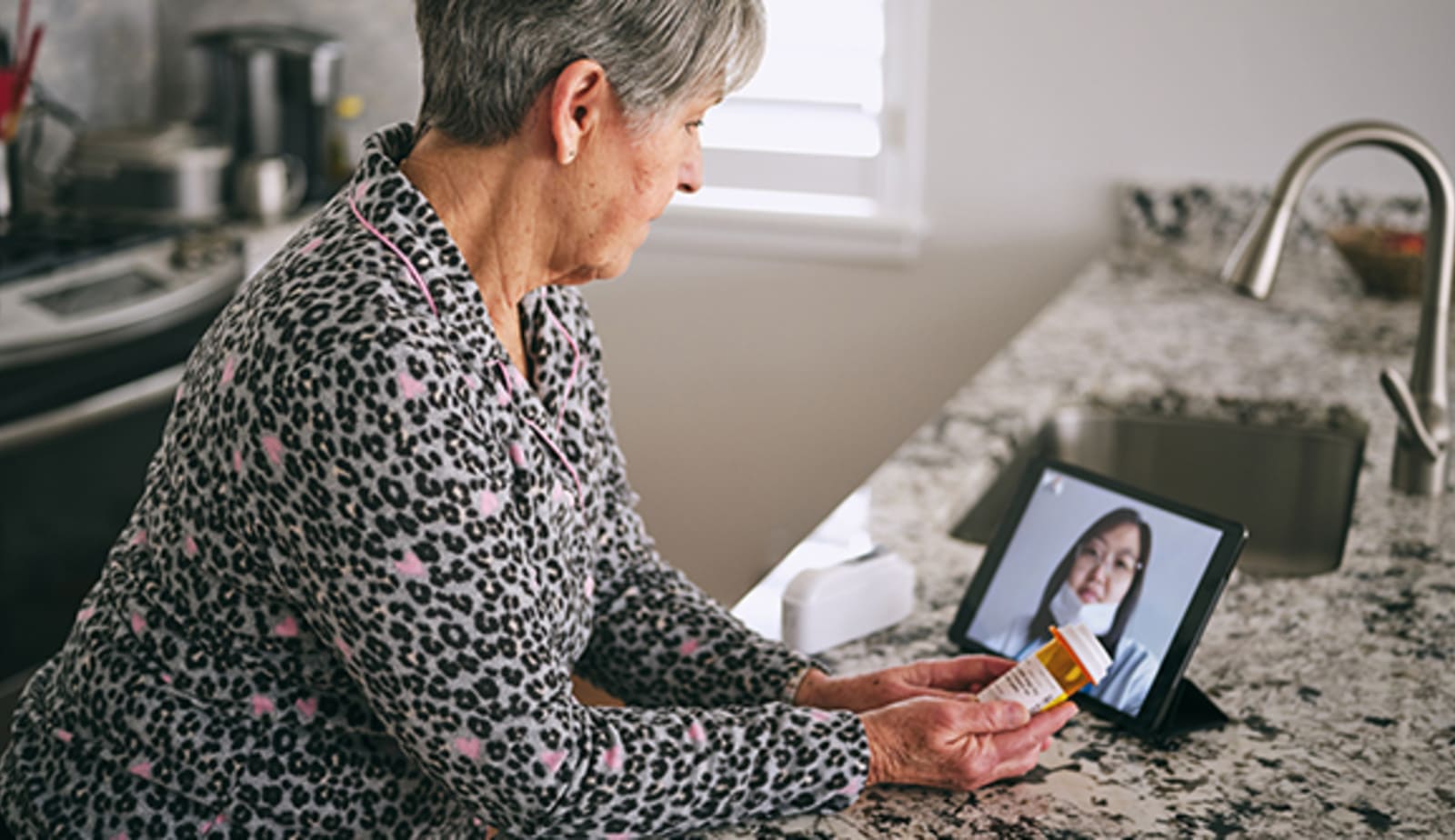 senior aged woman in her kitchen, using a tablet for a virtual appointment with a doctor