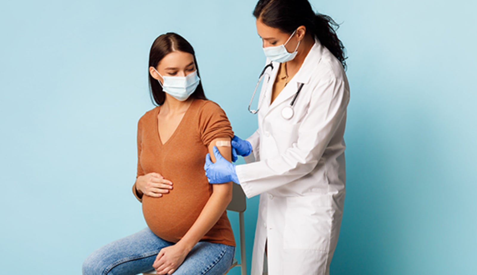 Masked nurse vaccinating pregnant female patient and sticking bandage on her arm