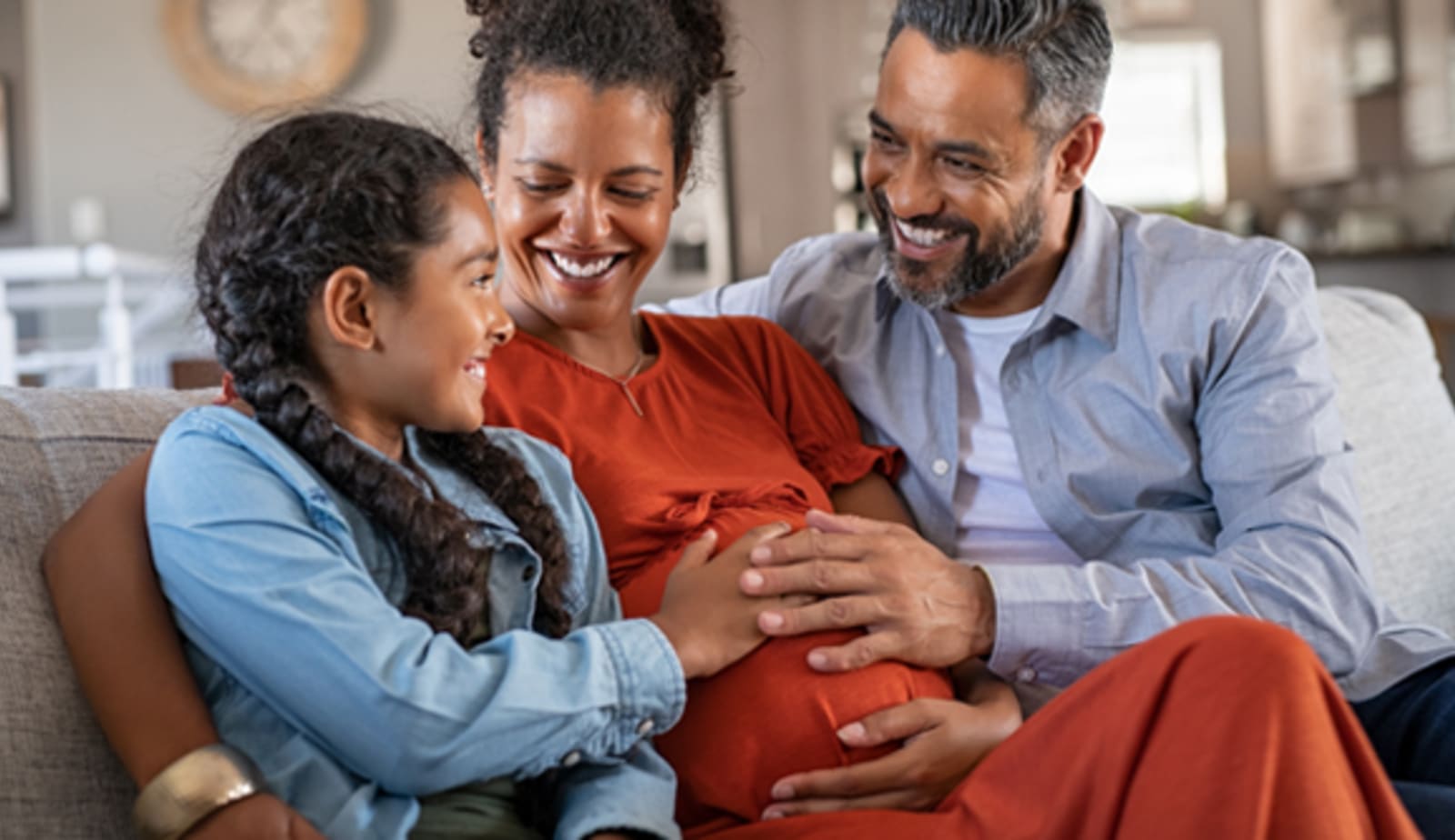 pregnant mother sitting on the couch with partner and daughter feeling her bump
