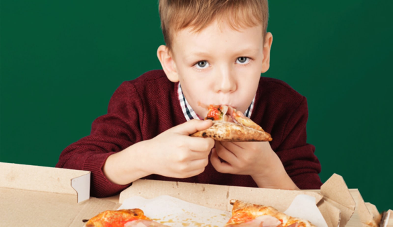 A young boy is staring into the camera as he shoves a cheesy slice of pizza into his mouth