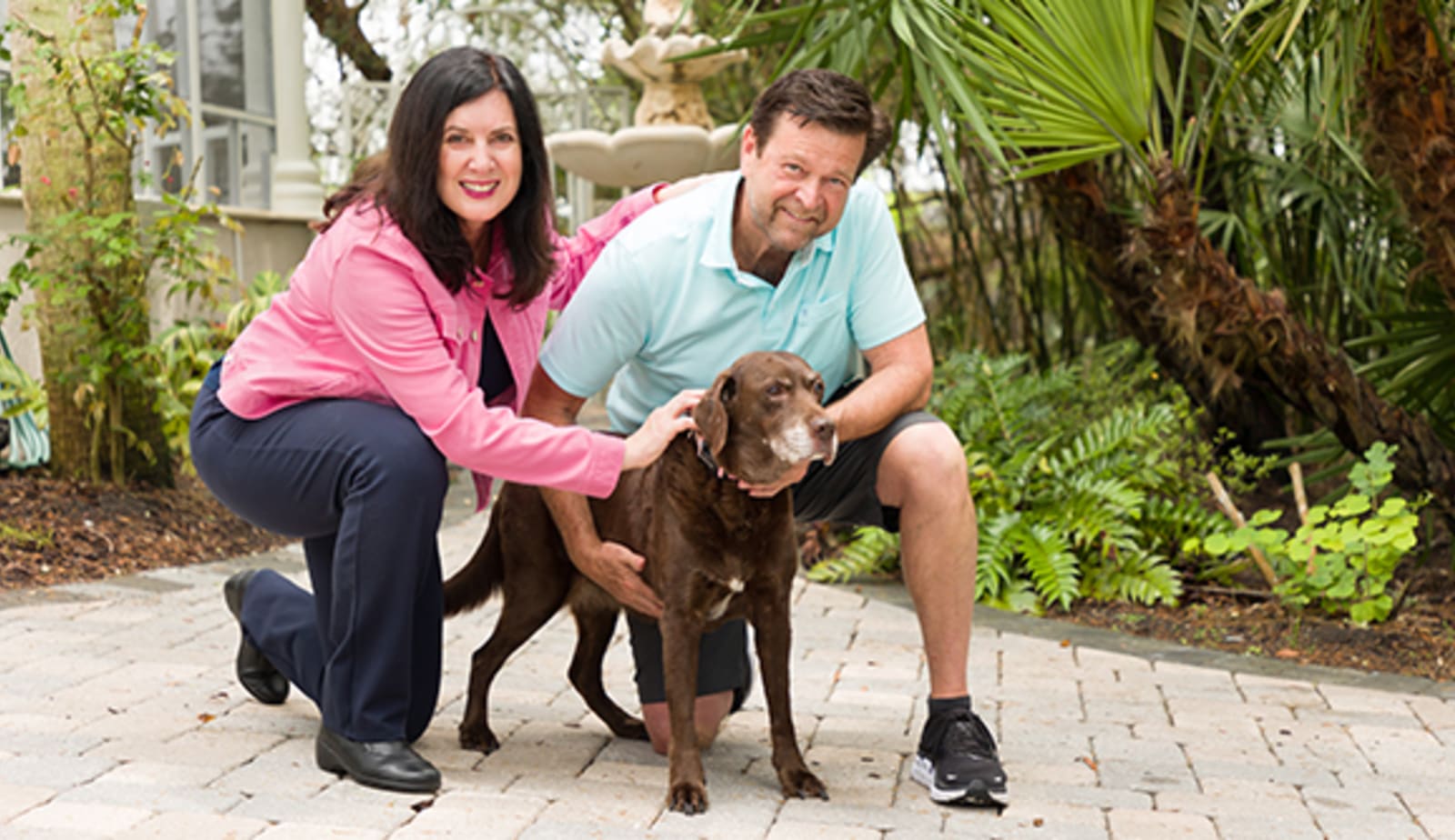 Man in pink jacket smiles on the left. Her fiance is on the right. They are crouched down around their cute chocolate lab.