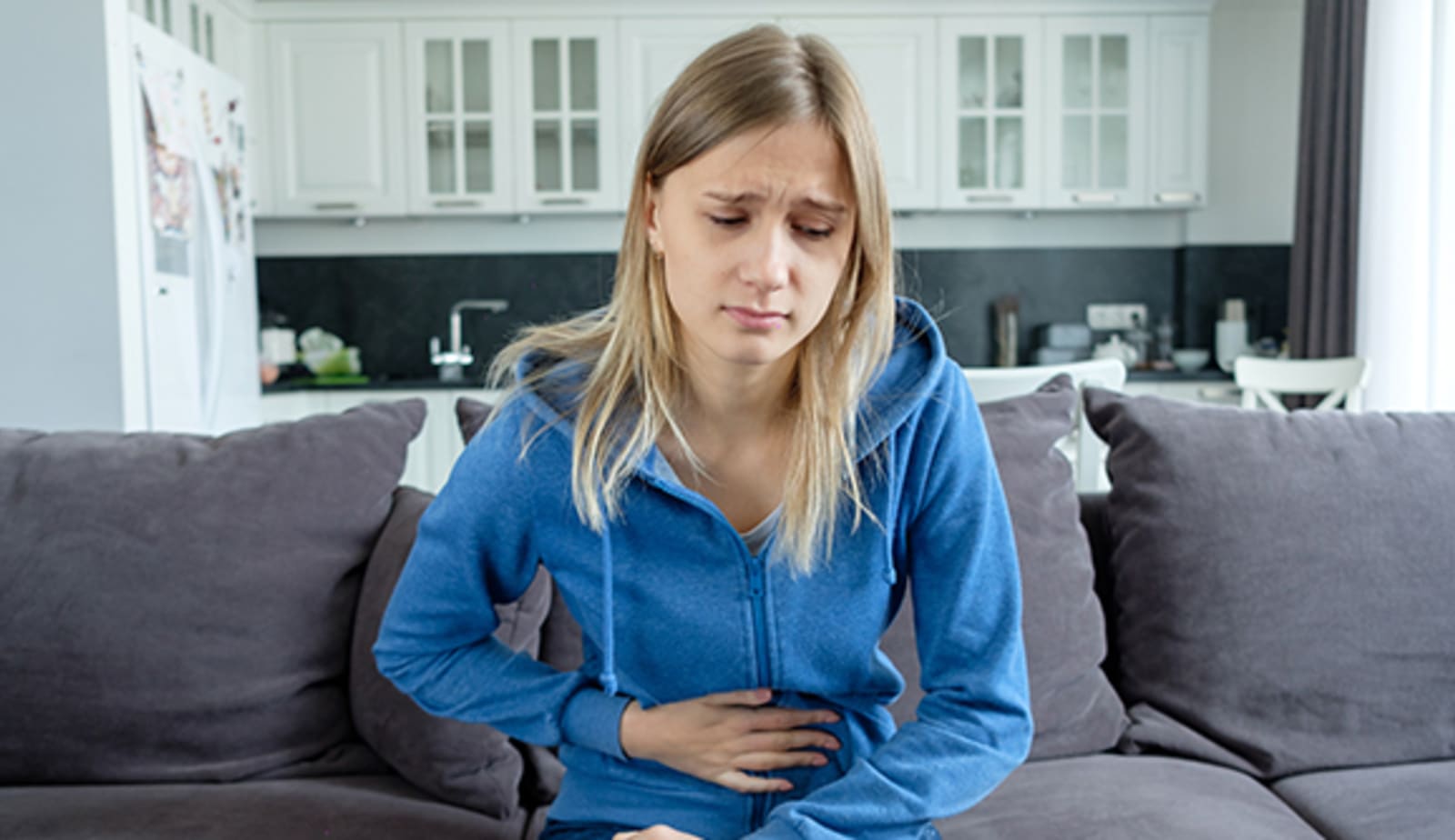 teenage girl holds her hand on a sore stomach while sitting on a couch