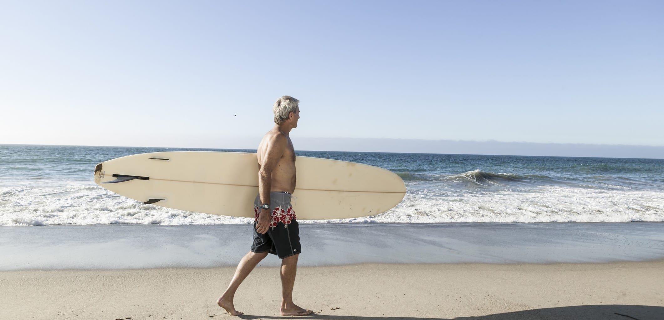 A man with gray hair walks along a beach carrying a surf board