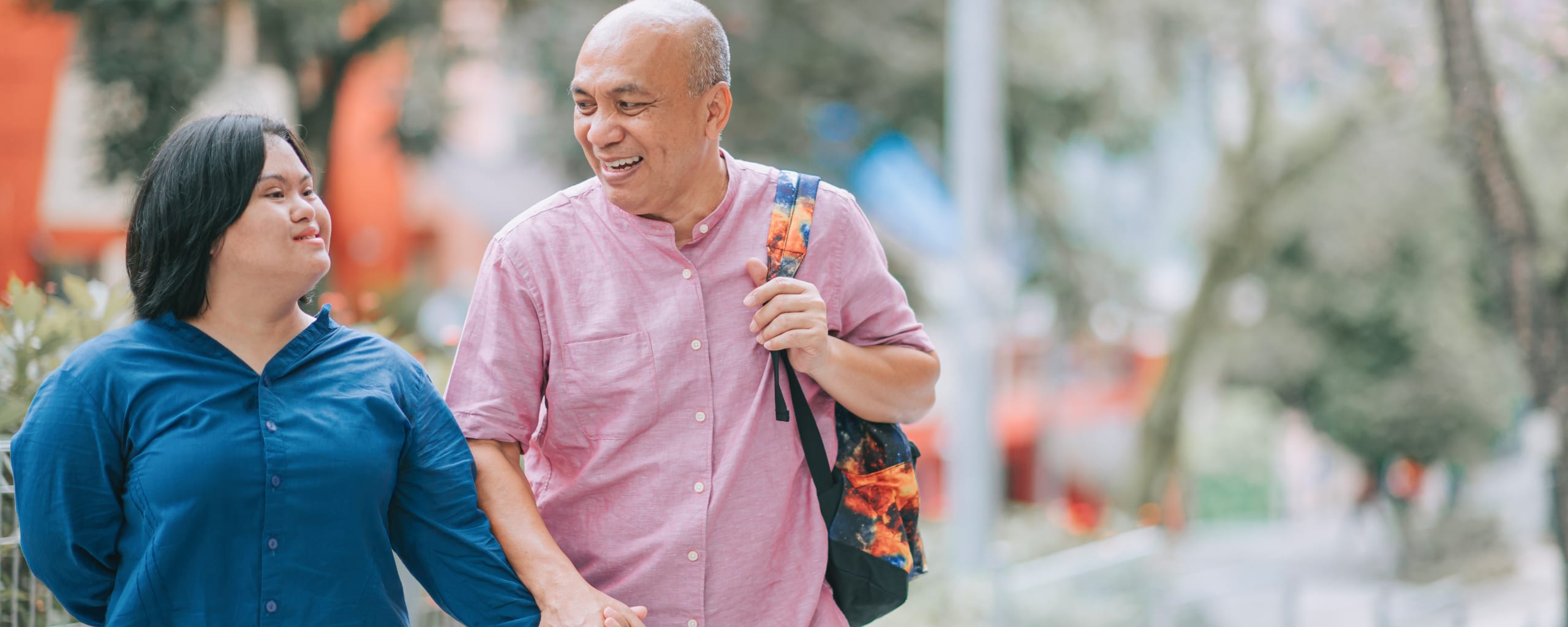 A young woman with Down syndrome holds the hand of an older man while walking down the street, both are smiling