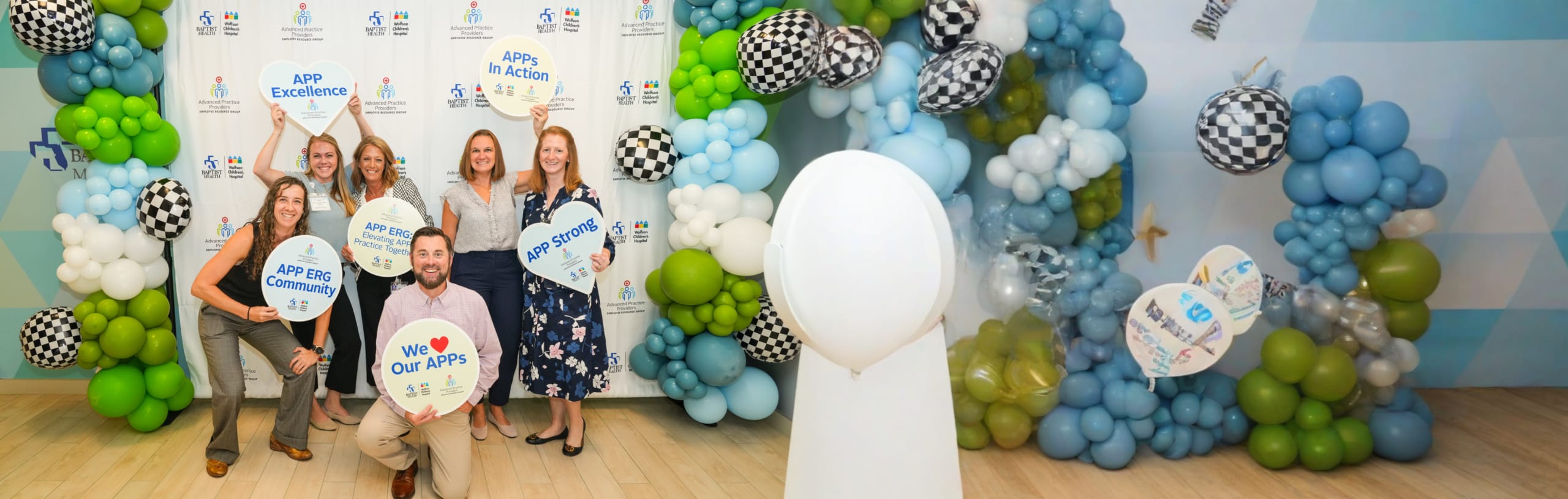 a group of smiling advanced practice providers at a Baptist Health event standing in front of festive balloons holding signs that say "APP Excellence," "We love our APPs," and "APP Strong"